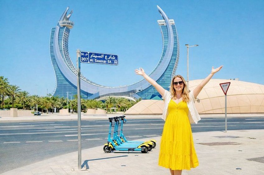 A woman in a yellow dress standing with arms outstretched, a blue scooter, and a distinctive modern building in the background. 1