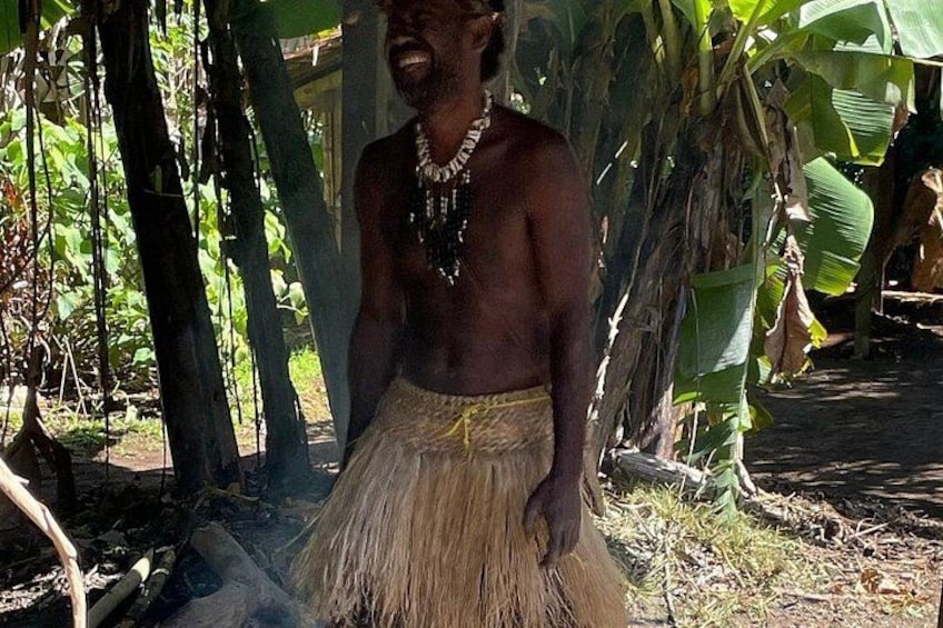 A person standing outdoors with a thatched skirt, surrounded by trees and a banana plant. 3