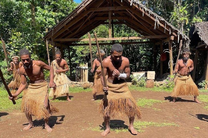 People in traditional attire performing a dance in an outdoor setting with a wooden structure in the background. 1