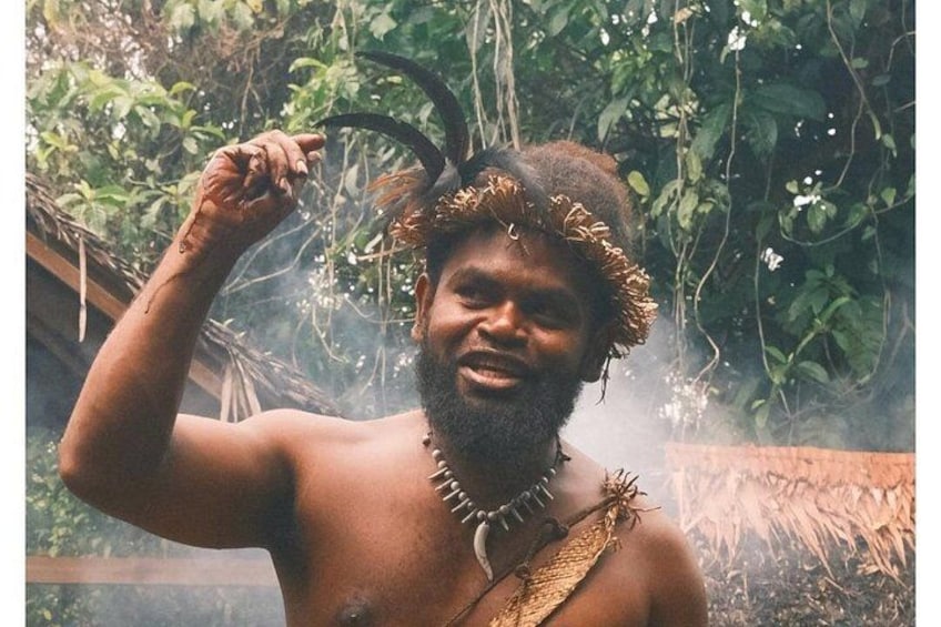 A person with a headdress in a natural setting with greenery and a structure in the background. 5