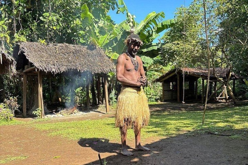 A person in traditional attire standing in a rural setting with thatched-roof huts and lush greenery. 4