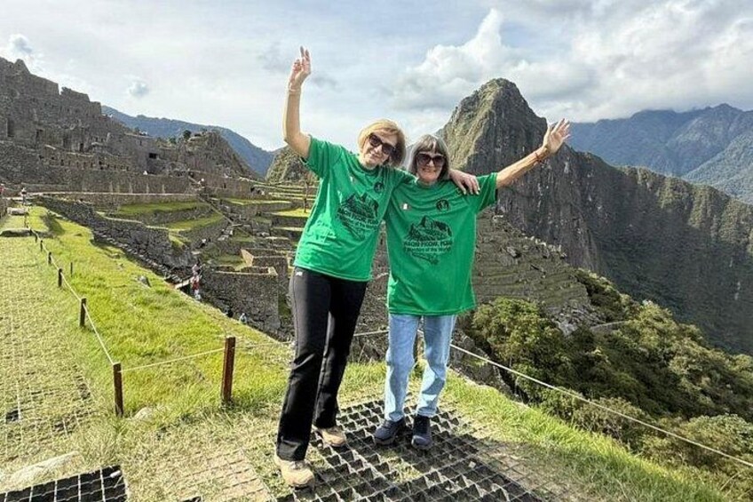 Dos personas de pie en una plataforma, con los brazos levantados, observando un paisaje montañoso con campos en terrazas. 1