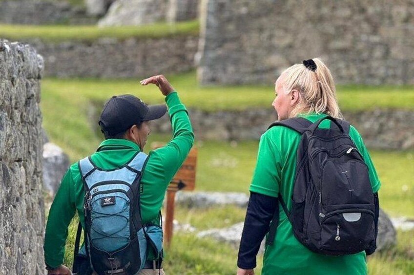Dos personas caminando con mochilas en un área de hierba con estructuras de piedra. 2