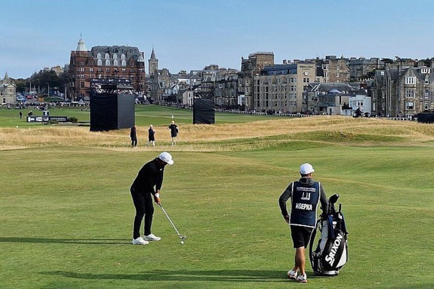 Ein Golfplatz mit Spielern und Zuschauern, im Hintergrund eine Stadtansicht und ein klarer Himmel. 2