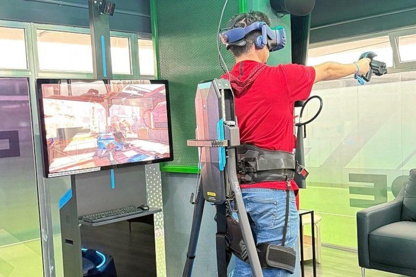 Una persona con camisa roja y jeans azules usando un casco de realidad virtual y sosteniendo un control, en una habitación con una pantalla grande que muestra un juego. 3