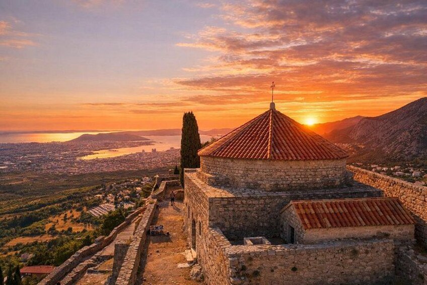 A historical stone building with a red-tiled roof, overlooking a valley and a body of water during sunset. 4