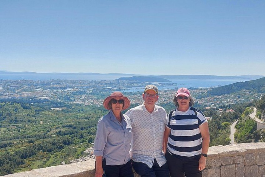 Three people standing on a stone wall overlooking a coastal landscape with greenery and a clear sky. 5
