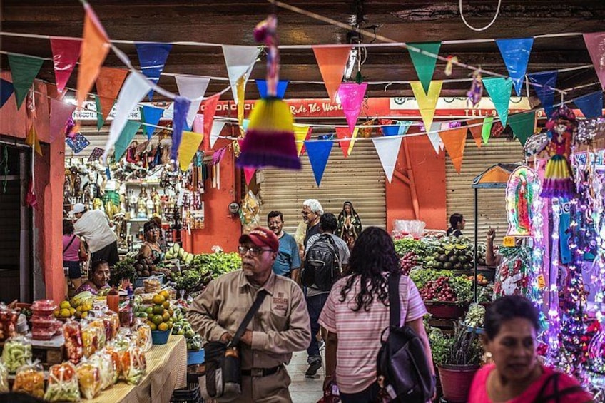 Un mercato vivace con bandiere colorate, diverse bancarelle che vendono frutta e verdura, e persone che fanno acquisti. 3