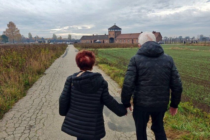 Two people walking on a paved path, holding hands, with a building and fields in the background. 3