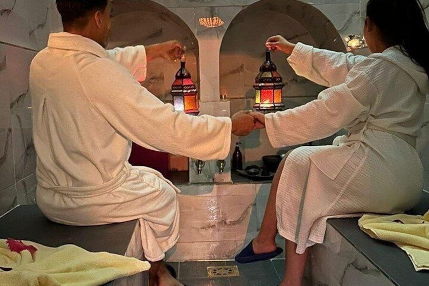 Two people in white robes sitting in a stone-walled room with a central fountain and two lit lanterns. 1