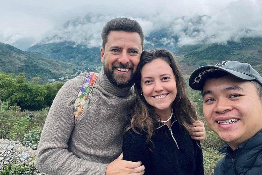 Three people standing on a mountain with a cloudy sky and greenery in the background. 3