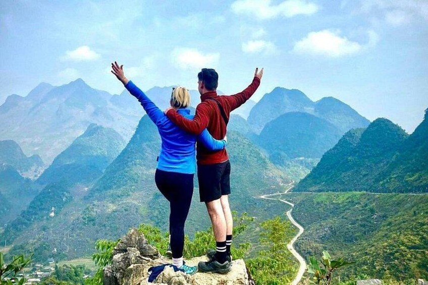 Two people standing on a rocky outcrop with arms raised, overlooking a valley with a winding road and mountains in the background. 2