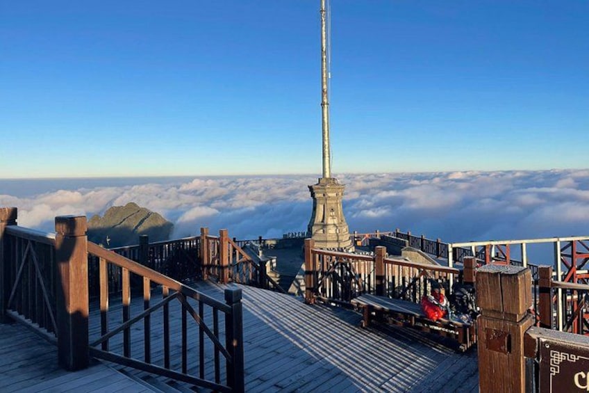 A wooden observation deck with a metal fence, a tall monument, and a mountain range above a cloud layer. 1