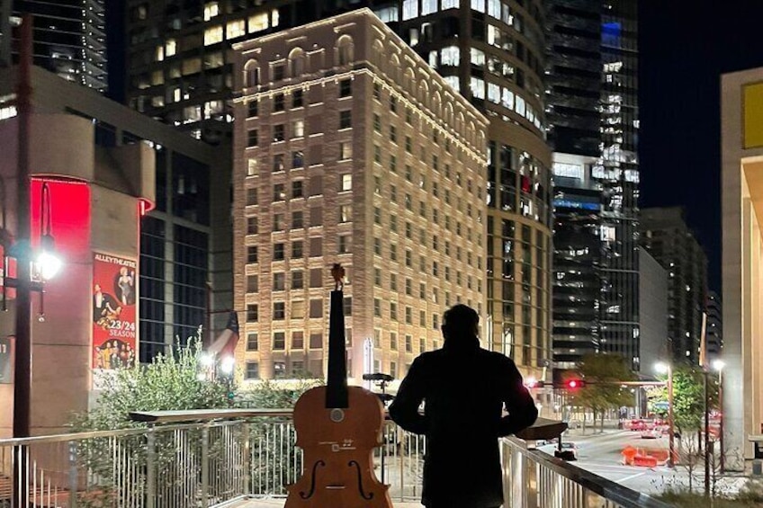 A silhouette of a person standing on a balcony with a guitar, overlooking a cityscape at night. 5
