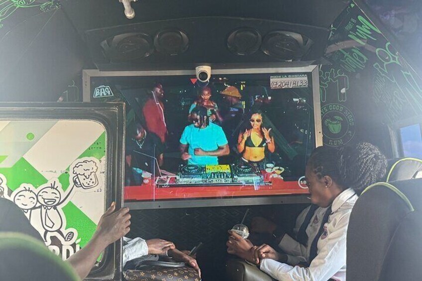 Een businterieur met een groot scherm waarop een DJ-optreden te zien is. 5