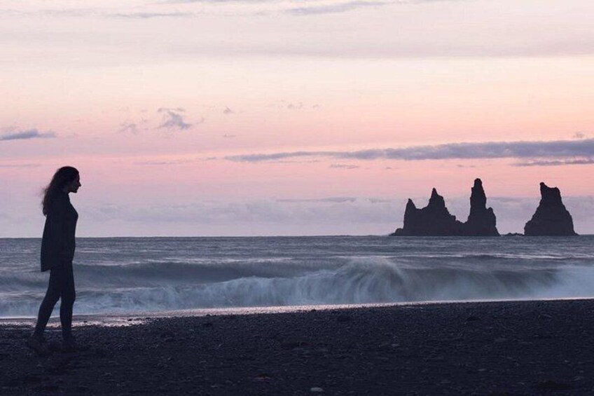 A silhouette of a person standing on a beach with waves crashing in the background and rock formations in the distance. 4