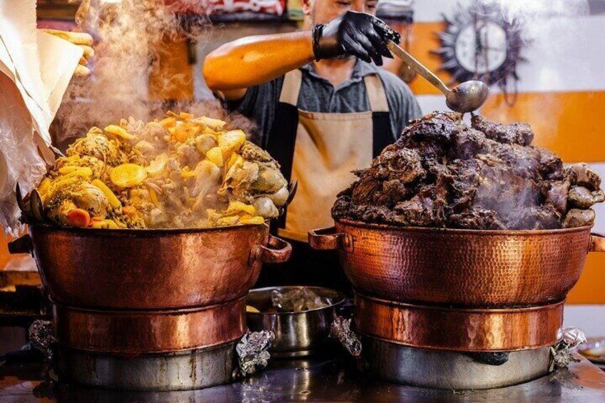 Deux grands récipients en cuivre sont posés sur une cuisinière : l’un contient un mélange de viande et de légumes qui mijote, et l’autre renferme un liquide sombre et bouillonnant. 1
