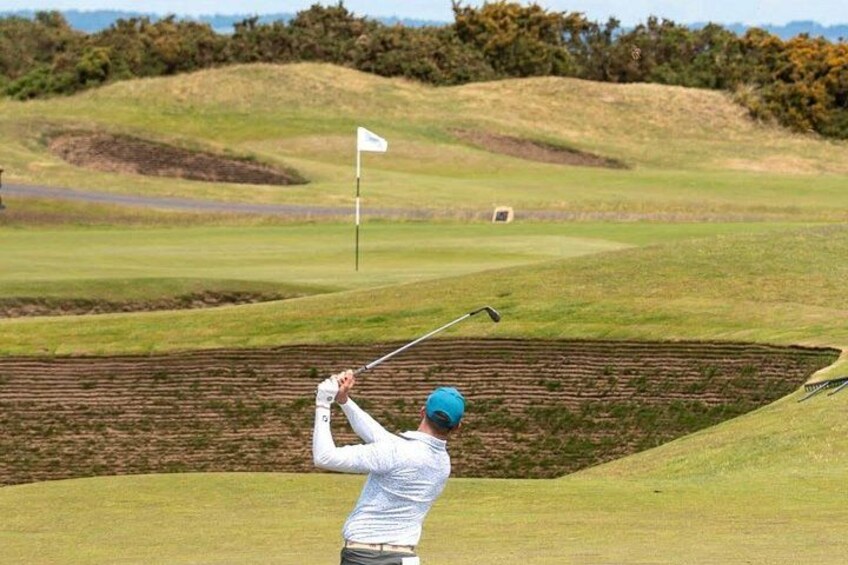 Ein Golfer in weißem Hemd und blauer Mütze bereitet sich auf dem Golfplatz vor, einen Ball zu schlagen, im Hintergrund weht eine Flagge. 3