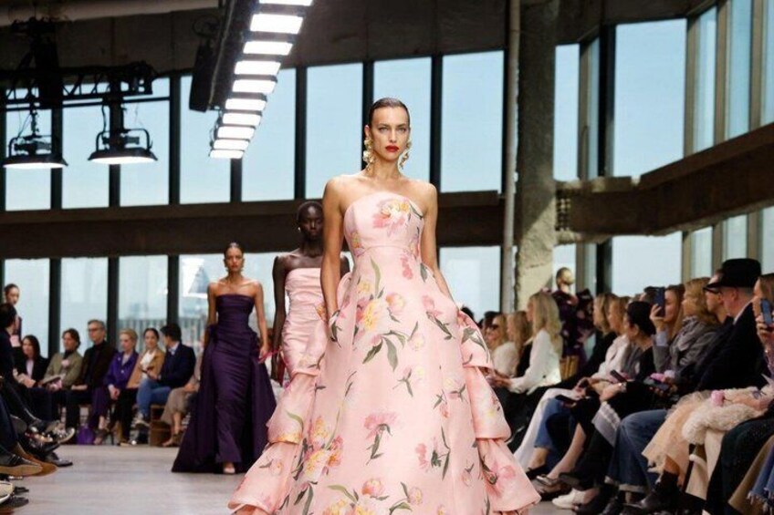 En catwalk där modeller visar upp klänningar, med åskådare sittande på båda sidor, och ett stort fönster i bakgrunden. 1