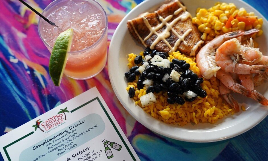 A plate of food with rice, black beans, shrimp, and grilled meat, alongside a tropical drink with a lime wedge. 3