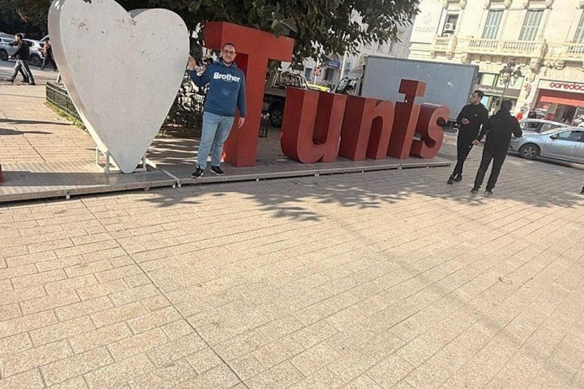 Ein großes, herzförmiges Schild mit dem Wort 'Tunis' und ein Mann in einem blauen Hemd, der daneben steht. 5