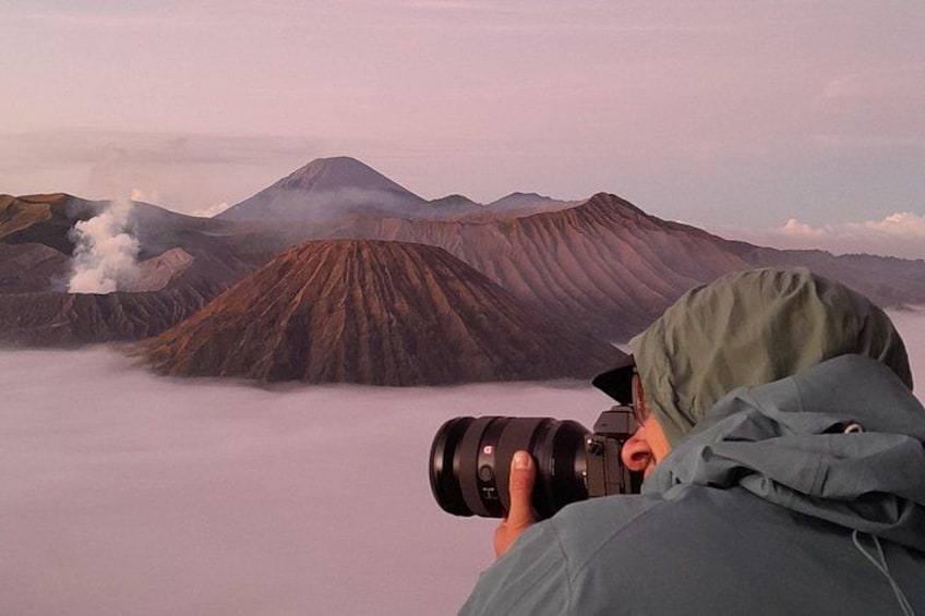 A person taking a photograph of a volcanic landscape with multiple peaks and smoke. 5