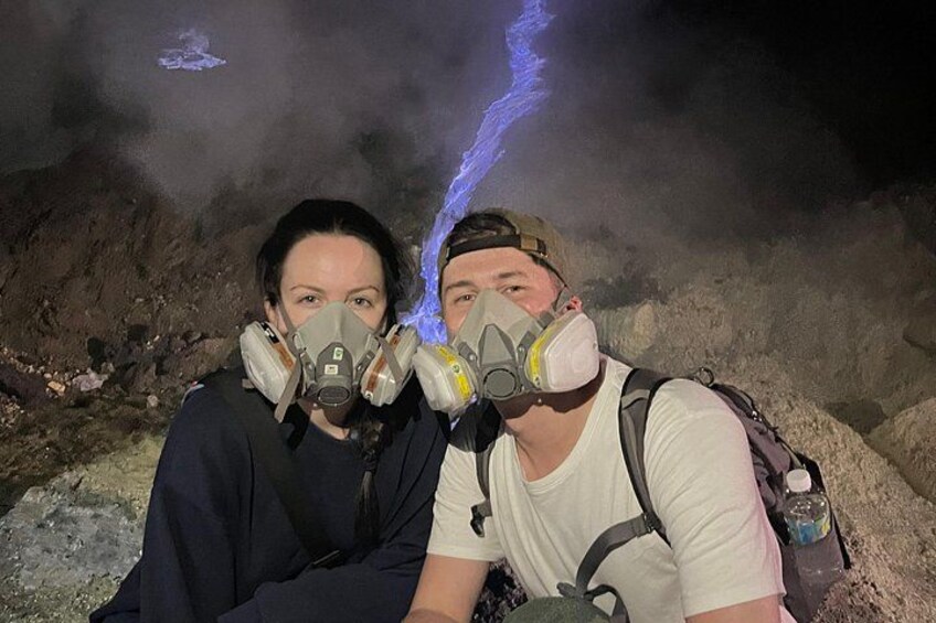 Two people wearing gas masks sitting in a rocky area with a blue light in the background. 3