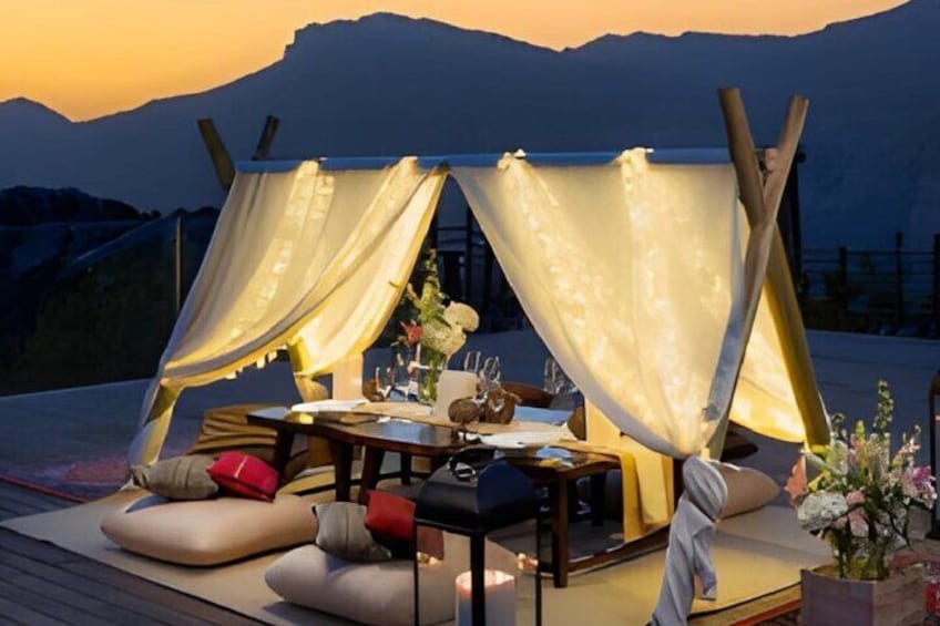 A cozy outdoor dining setup with a table and chairs, draped with white curtains, set against a mountainous backdrop during sunset. 2