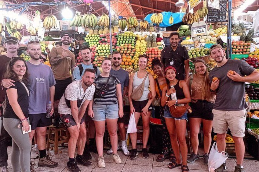 Un groupe de personnes posant devant un marché aux fruits et légumes, où sont exposés divers fruits et légumes. 1
