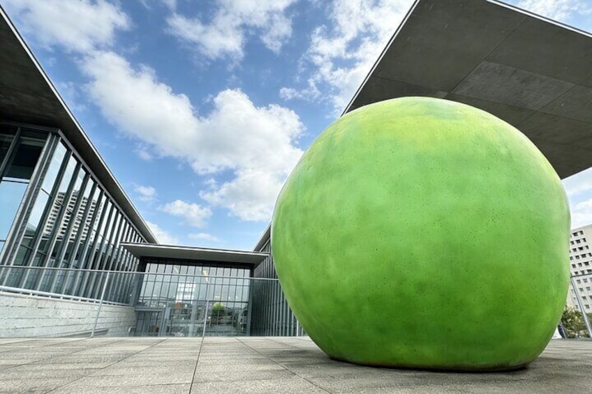 Una gran escultura esférica verde en una plaza urbana con edificios modernos al fondo. 1