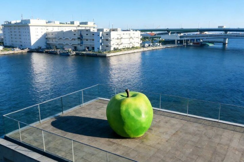 A large green apple sculpture on a waterfront platform with buildings and a bridge in the background. 3