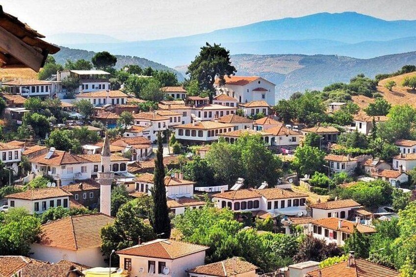 A hillside village with terracotta-roofed houses, a prominent church tower, and a backdrop of mountains. 1