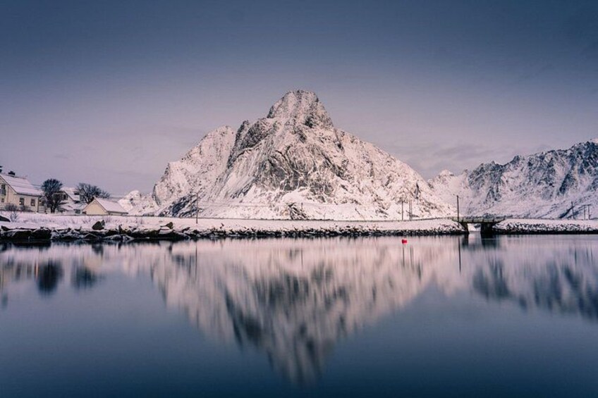 A snow-covered mountain reflected in a calm lake, with a bridge in the foreground and houses on the left. 2
