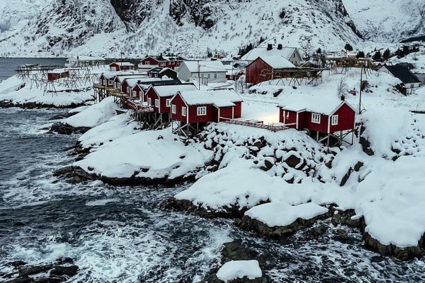Ein Küstendorf mit roten Häusern auf Stelzen, umgeben von schneebedeckten Bergen und felsigen Ufern. 3