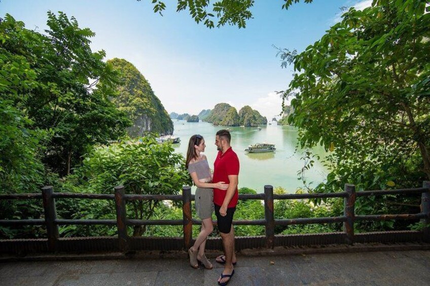 A couple standing by a river with a boat in the distance, surrounded by lush greenery and a wooden railing. 1