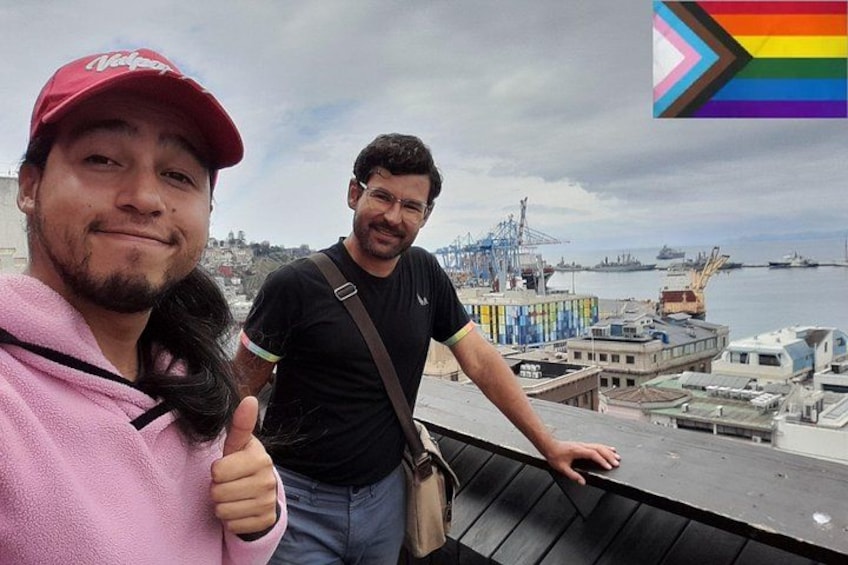 Two people standing on a rooftop with a view of a harbor and ships in the background. 1
