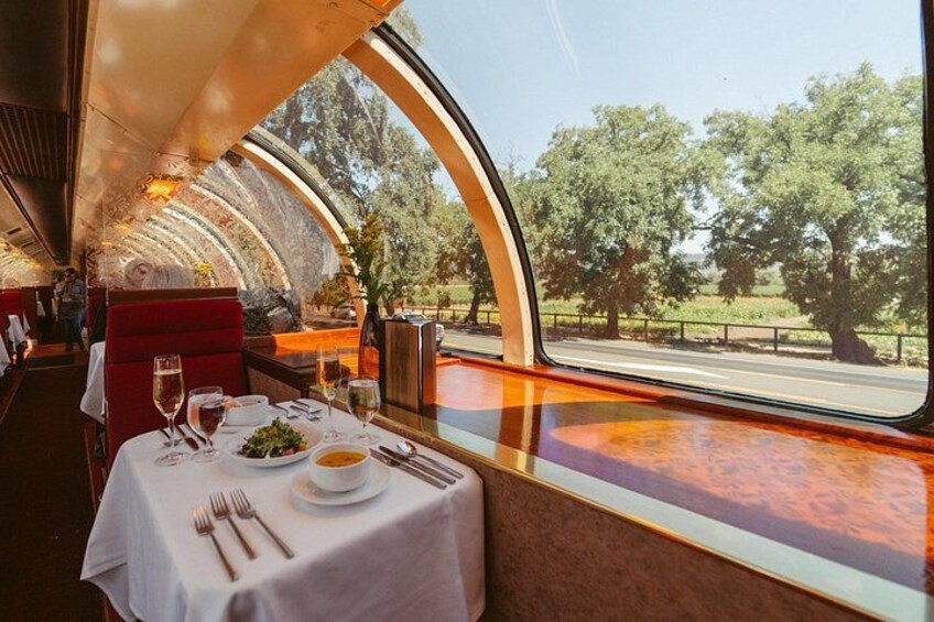 A dining car with a table set for two, a meal, and a view of trees and a road outside. 3