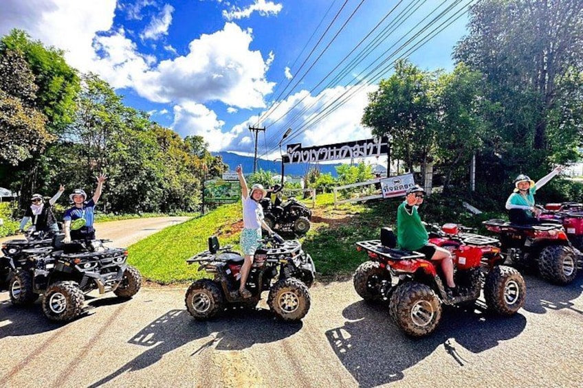 People on ATVs on a road with greenery and a sign in the background. 1