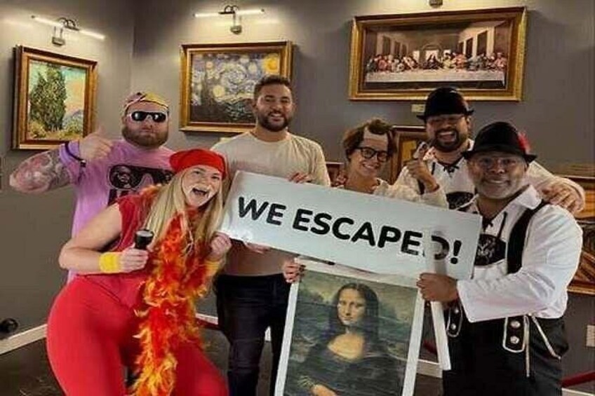 Eine Gruppe von Menschen in einem Raum, an den Wänden hängen Gemälde, eine Person hält ein Schild mit der Aufschrift 'WE ESCAPE!'. 3