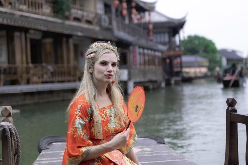 A person in traditional attire holding a fan, seated on a boat in a river with traditional architecture in the background. 4