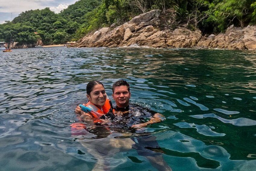Two people in a body of water with a rocky shoreline and dense greenery in the background. 3