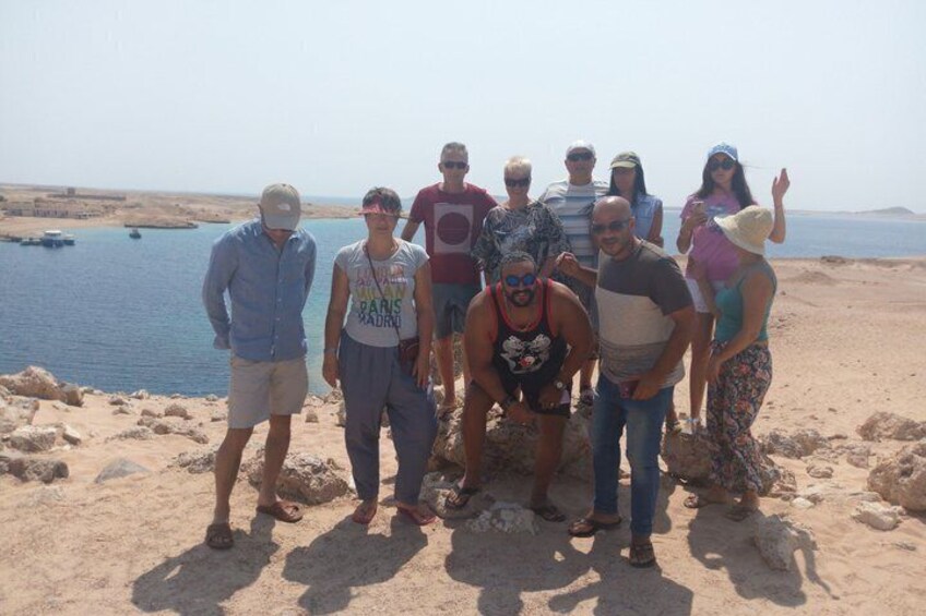 En gruppe mennesker står på en sandstrand med vand og klippelandskab i baggrunden. 4