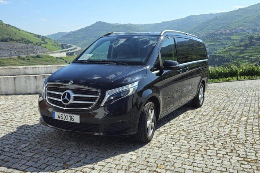 A black Mercedes-Benz minivan parked on a cobblestone surface with a scenic landscape in the background. 1