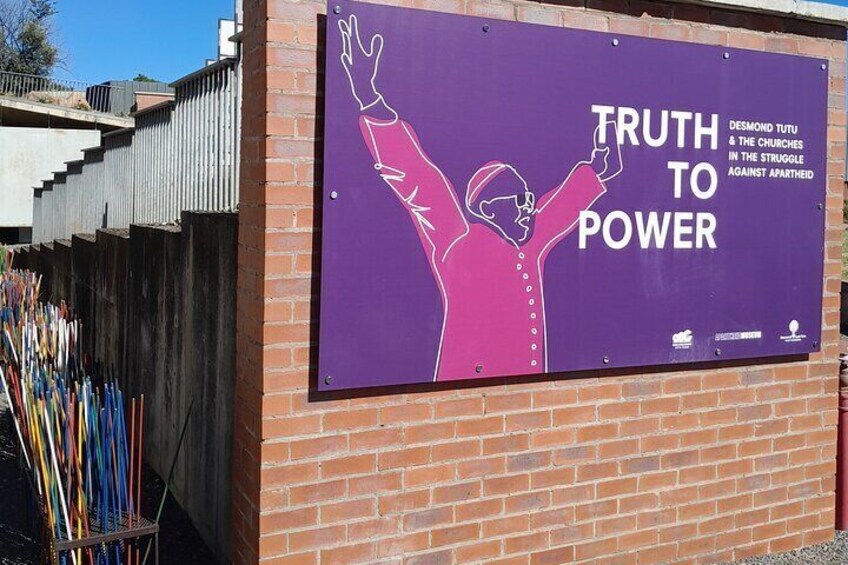 En lila skylt med en person som lyfter armarna och texten ”SÄG SANNINGEN TILL MAKTEN” och ”DESMOND TUTU OCH KYRKORNA I KAMPEN MOT APARTHEID”. 1