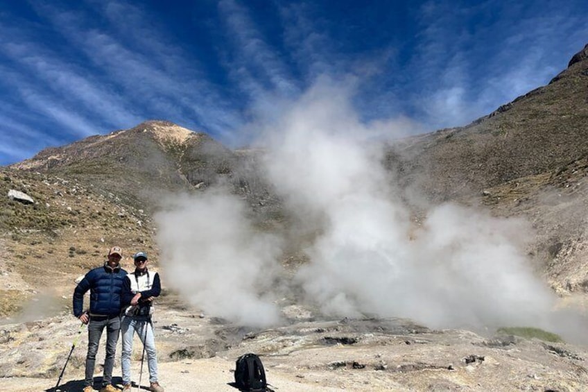 Two people standing near a geothermal area with steam rising from the ground. 1