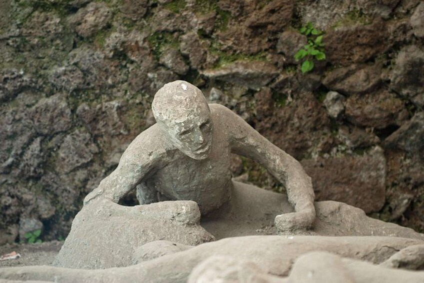 A stone sculpture of a human figure in a crouched position, possibly a representation of a historical or cultural scene, set against a rocky background. 3