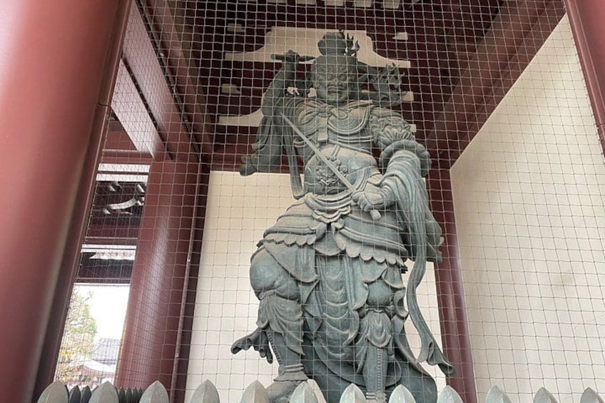 Un'imponente statua di una figura in abiti tradizionali, forse una divinità, eretta in un tempio o santuario con tetto in tegole e una recinzione in primo piano. 4