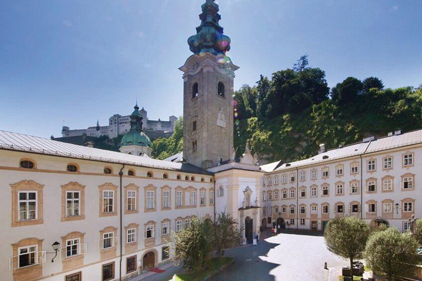 A historic building with a tower, a courtyard, and a hill with trees in the background. 1