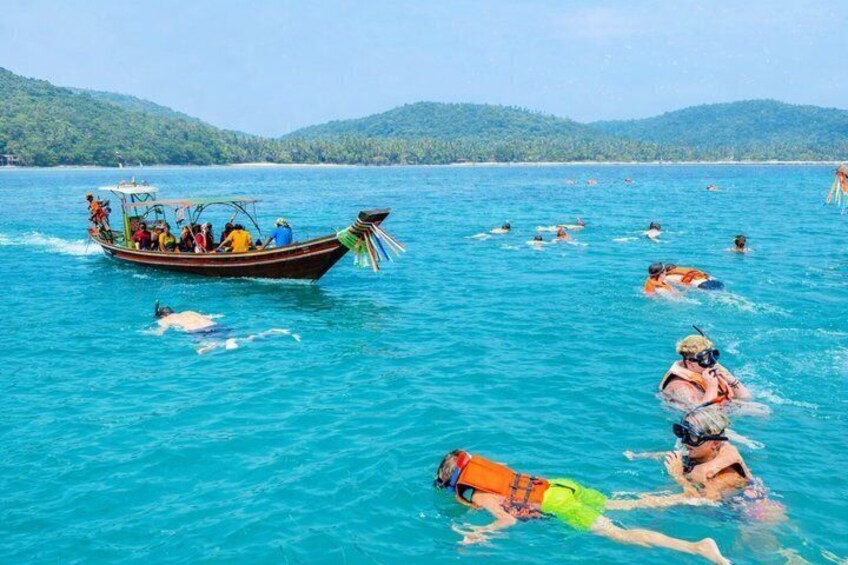 Un bote transporta personas sobre un mar turquesa, con nadadores en el agua y un paisaje montañoso al fondo. 1
