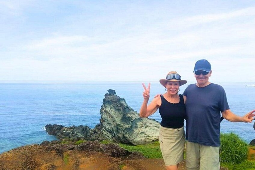 A rocky coastline with a prominent rock formation, a person making a peace sign, and another person standing beside them. 3